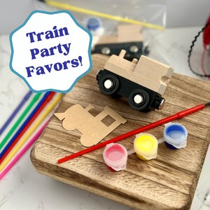 May include: Wooden train party favors set with paint, brushes, and colorful straws. The set includes a wooden train, a train cutout, and small paint pots in pink, yellow, and blue. The image also includes the text "Train Party Favors!"