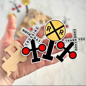 May include: A collection of train-themed stickers and cutouts. Includes a yellow and black railroad crossing sign, crossing signals with red circles, and a train-shaped cutout. Text on the stickers reads "BIRTHDAY CROSSING", "THANK YOU", and "CHOO CHOO".