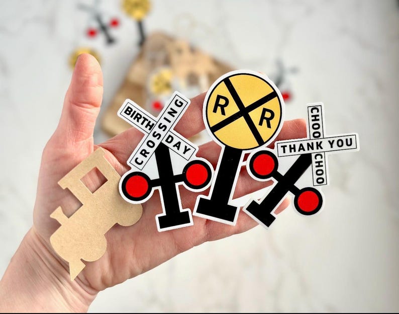 May include: A collection of train-themed stickers and cutouts. Includes a yellow and black railroad crossing sign, crossing signals with red circles, and a train-shaped cutout. Text on the stickers reads "BIRTHDAY CROSSING", "THANK YOU", and "CHOO CHOO".
