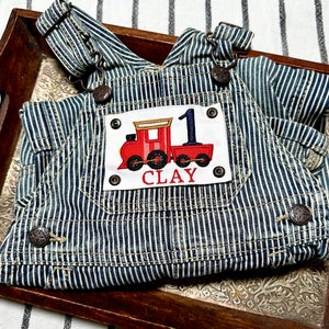 Puede incluir: Peto vaquero a rayas azules y blancas con un aplique de tren rojo. El aplique presenta el número 1 y el nombre "CLAY". El peto tiene botones metálicos y tirantes ajustables.