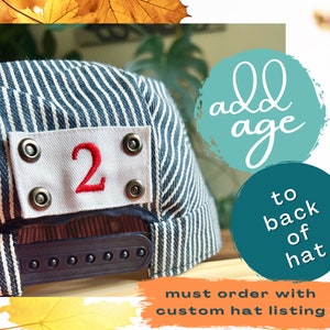 Puede incluir: Una gorra de béisbol a rayas azules y blancas con un parche blanco en la parte delantera que dice "2" en rojo. El texto "add age to back of hat must order with custom hat listing" está escrito en la imagen.