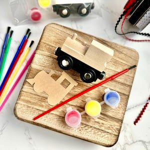 Puede incluir: Un kit de manualidades de tren de madera con un tren de madera, pinceles, botes de pintura y cinta. El tren es de madera natural con ruedas negras. Los colores de pintura incluyen rojo, amarillo y azul. El kit está sobre una tabla de cortar de madera.