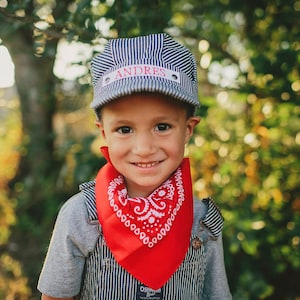 May include: A young boy wearing a blue and white striped engineer's hat with the name "ANDRES" on it. He is also wearing a red bandana with a white pattern around his neck.