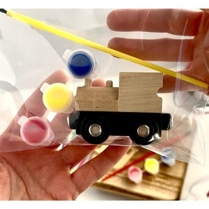 May include: A wooden train model kit with paint pots in pink, yellow, and blue, and a yellow paintbrush. The train is black and natural wood, packaged in a clear plastic bag. A fun craft project for kids.
