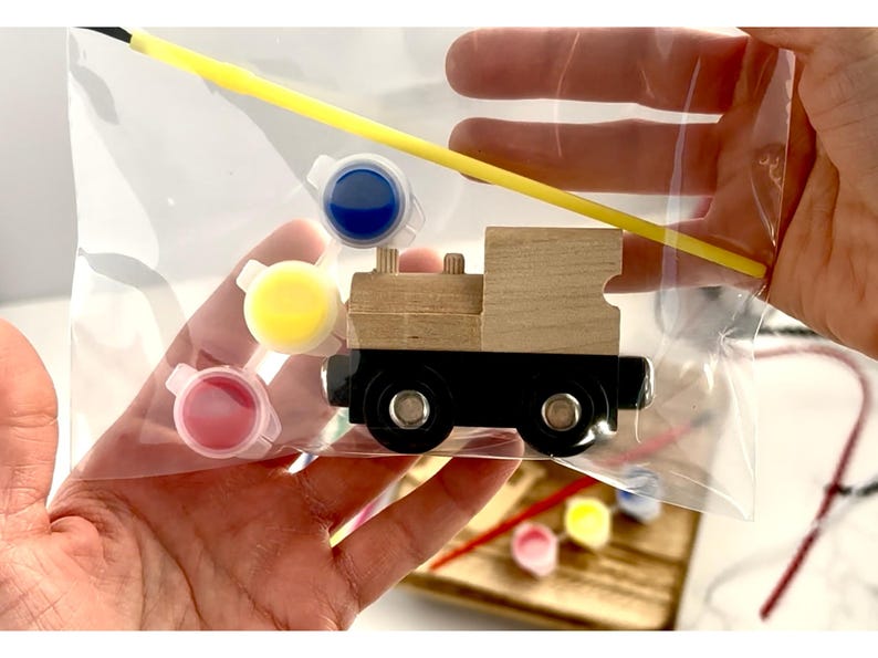 May include: A wooden train model kit with paint pots in pink, yellow, and blue, and a yellow paintbrush. The train is black and natural wood, packaged in a clear plastic bag. A fun craft project for kids.