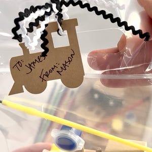 May include: A clear plastic bag containing a craft kit. A brown paper train-shaped tag is attached with black and white ribbon. The tag reads "To: James From: Mason". A yellow paintbrush and a small container of blue paint are visible.