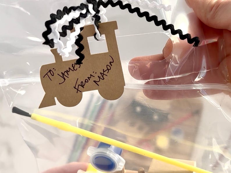May include: A clear plastic bag containing a craft kit. A brown paper train-shaped tag is attached with black and white ribbon. The tag reads "To: James From: Mason". A yellow paintbrush and a small container of blue paint are visible.
