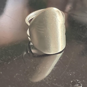 May include: A silver-colored ring with a large, oval-shaped face. The ring has a smooth, metallic finish and a simple band. The face of the ring has subtle, textured details. The ring is displayed on a reflective surface.