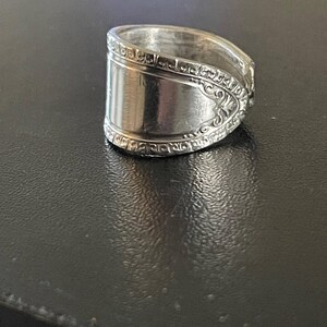 Puede incluir: Un anillo de plata hecho de una cuchara, con una banda ancha y un patrón ornamentado en los bordes. El anillo tiene una superficie pulida y reflectante. El diseño muestra la cubertería reutilizada.