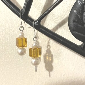May include: A pair of earrings with a silver hook, a white pearl, and a yellow glass cube. The earrings are hanging from a black metal object.
