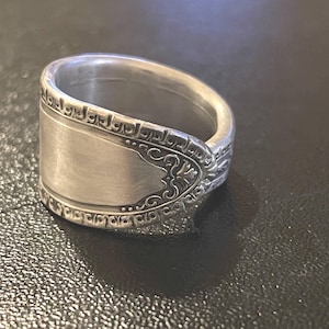 May include: A silver ring crafted from a spoon, featuring a rectangular face with ornate scrollwork and a decorative border. The ring has a polished finish and a vintage aesthetic.