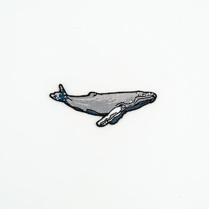May include: An embroidered patch featuring a humpback whale in shades of grey, blue, and white. The whale is depicted in a side profile view with a textured surface, detailed stitching, and a dark outline, showcasing its fins and tail fluke.