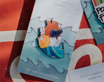 May include: Enamel pin with a heart design and ocean waves in orange, blue, and white. The pin is on a card that reads "WINGS HAWAII ENAMEL PIN" and "FOR REAL LIFE MERMAIDS". Other cards with pins are visible.