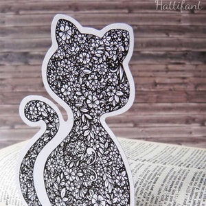 Op de afbeelding: Een zwart-witte bladwijzer met een katten-silhouet dat is gevuld met ingewikkelde bloemmotieven. De bladwijzer is gelabeld met "Flower Cat Bookmarks".
