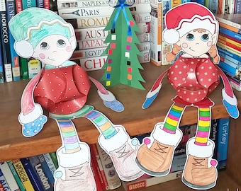 May include: Two paper elf ornaments, one with a green hat and red sweater, the other with a red hat and red sweater, both with rainbow striped legs and brown boots. The elves are sitting on a bookshelf with books behind them.