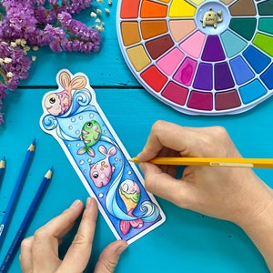 May include: A colourful bookmark featuring a design of three fish swimming in a blue wave. The bookmark is on a blue surface with a hand holding a yellow pencil.