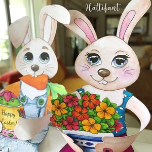 May include: Two paper Easter bunny figures. One bunny holds a carrot and an Easter egg with the text "Happy Easter!". The other bunny holds a bouquet of colorful flowers. The bunnies have white fur, pink ears, and blue eyes. The image has the text "Hattifant".