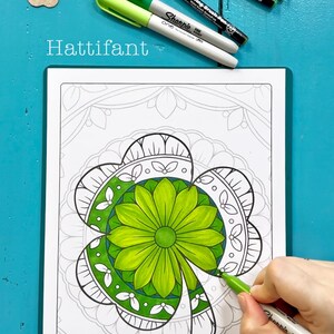 May include: A close-up of a colouring page featuring a detailed mandala design of a four-leaf clover. The clover is partially coloured in shades of green, and a hand is holding a green marker to colour the rest of the design. The text "Hattifant Design" is visible at the bottom of the page.