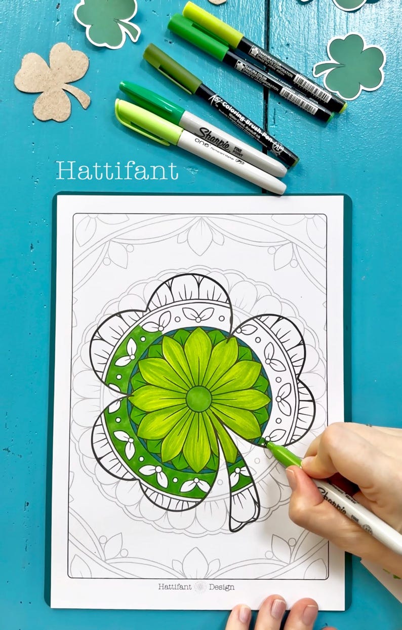 May include: A close-up of a colouring page featuring a detailed mandala design of a four-leaf clover. The clover is partially coloured in shades of green, and a hand is holding a green marker to colour the rest of the design. The text "Hattifant Design" is visible at the bottom of the page.