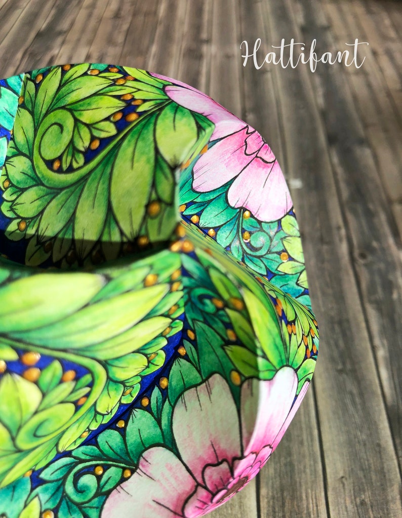Pode incluir: Close-up de um artesanato em papel com um design floral. O artesanato apresenta folhas verdes, flores rosa e detalhes dourados sobre um fundo azul. A palavra "Hattifant" est&aacute; escrita em branco.