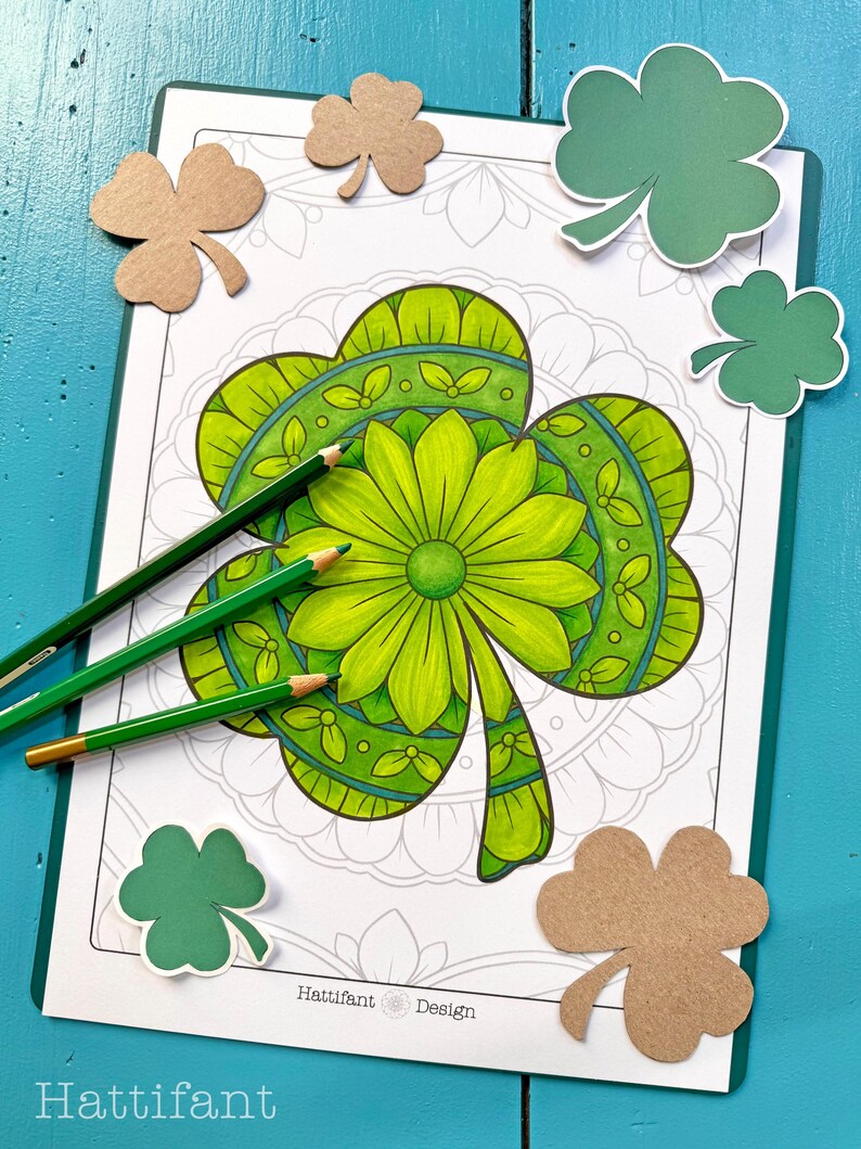 May include: A printable colouring page featuring a detailed shamrock design with green, blue and white colours. The shamrock is surrounded by a mandala pattern. The image includes the text "Hattifant Design".