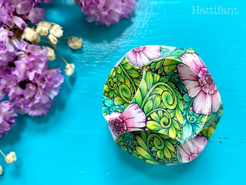 Pode incluir: Um artesanato em papel geom&eacute;trico colorido com um design floral. A obra de arte apresenta flores rosa e folhas verdes com detalhes dourados. O texto "Hattifant" est&aacute; no canto superior direito. O artesanato est&aacute; em uma superf&iacute;cie azul brilhante com flores roxas.