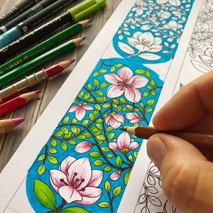 Pode incluir: Um marcador de p&aacute;gina parcialmente colorido com um design floral, apresentando flores rosa e folhas verdes sobre um fundo azul. L&aacute;pis de cor e marcadores s&atilde;o vis&iacute;veis, sugerindo uma atividade de colorir. A palavra "Hattifant" &eacute; vis&iacute;vel.