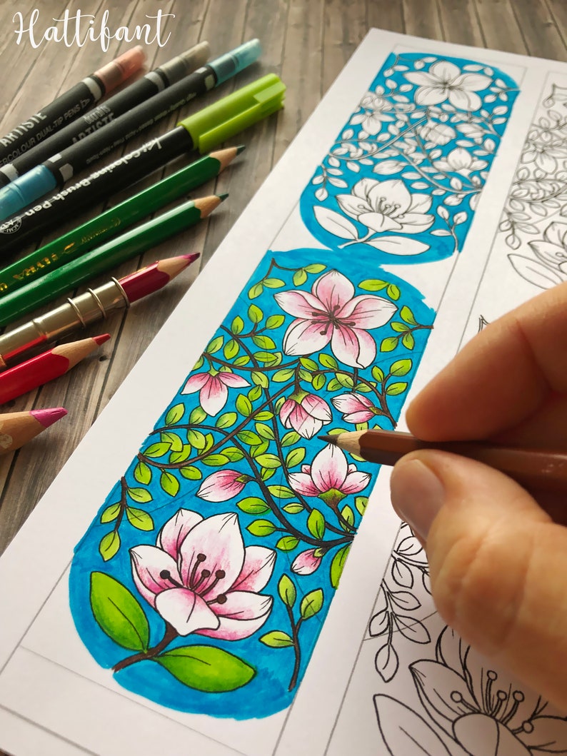 Pode incluir: Um marcador de p&aacute;gina parcialmente colorido com um design floral, apresentando flores rosa e folhas verdes sobre um fundo azul. L&aacute;pis de cor e marcadores s&atilde;o vis&iacute;veis, sugerindo uma atividade de colorir. A palavra "Hattifant" &eacute; vis&iacute;vel.