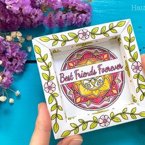 Pode incluir: Um quadro quadrado branco com uma borda de design floral. Dentro do quadro há um mandala colorido com o texto "Best Friends Forever".
