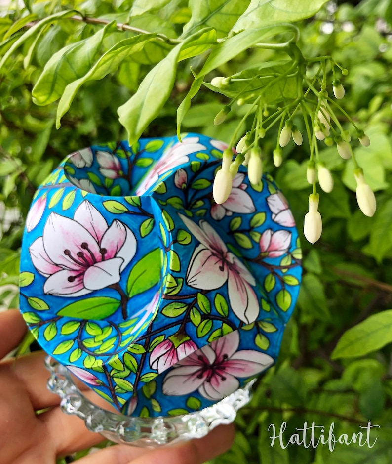 Pode incluir: Um artesanato de papel azul, possivelmente um vaso ou objeto decorativo, adornado com padr&otilde;es florais brancos e rosa e folhas verdes. Ele repousa sobre uma base de vidro transparente. O fundo &eacute; uma folhagem verde vibrante.
