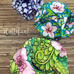 Pode incluir: Tr&ecirc;s chap&eacute;us de tecido coloridos com desenhos florais s&atilde;o exibidos em uma superf&iacute;cie de madeira. Os chap&eacute;us apresentam padr&otilde;es intrincados de flores e folhas em tons de azul, rosa e verde. A palavra "Hattifant" est&aacute; escrita em it&aacute;lico.