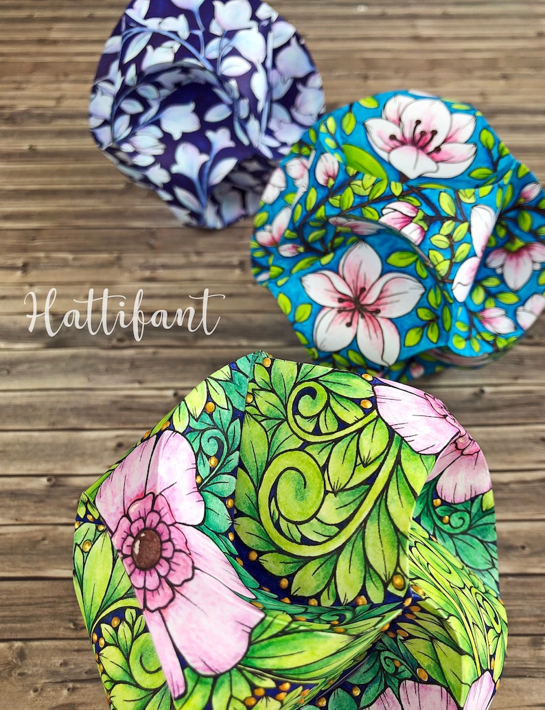 Pode incluir: Tr&ecirc;s chap&eacute;us de tecido coloridos com desenhos florais s&atilde;o exibidos em uma superf&iacute;cie de madeira. Os chap&eacute;us apresentam padr&otilde;es intrincados de flores e folhas em tons de azul, rosa e verde. A palavra "Hattifant" est&aacute; escrita em it&aacute;lico.