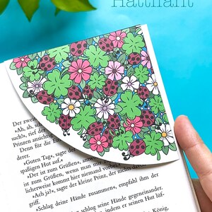 May include: A close-up of a book with a bookmark featuring a colorful floral design with ladybugs. The text on the page reads "Hattifant" and includes a German story about a prince.