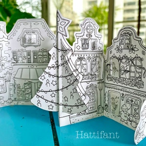 May include: A black and white printable paper craft of a Christmas village with a Christmas tree in the center. The village has four buildings with windows and doors. The buildings are decorated with Christmas lights and ornaments. The text "Hattifant" is printed at the bottom of the image.