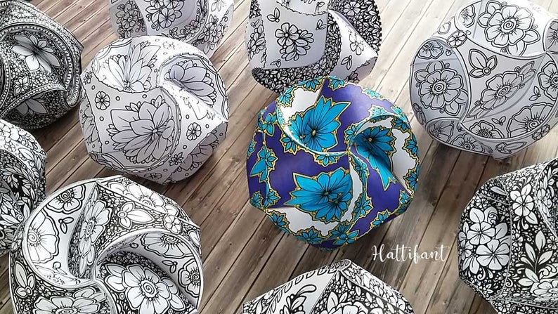 May include: A collection of eight origami paper balls with intricate floral designs. The balls are in various shades of white, black, blue, and gold. The balls are arranged on a wooden surface.