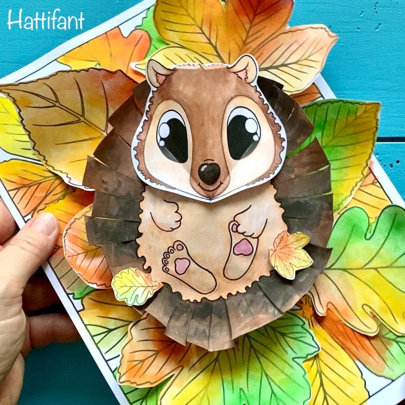 May include: A paper craft of a brown hedgehog with large eyes sitting in a nest of colorful fall leaves. The hedgehog is surrounded by leaves in shades of orange, yellow, and green.