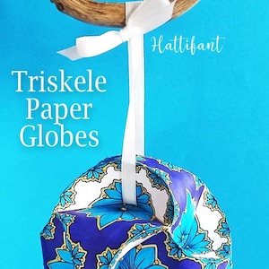 May include: A white ribbon hangs from a branch, with a blue and white paper globe ornament attached. The globe has a floral pattern and the text "Triskele Paper Globes" is written above it. The text "Hattifant" is written in the upper right corner.