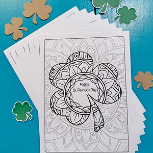 May include: A stack of blank white cards featuring a black and white printable design of a shamrock with the text "Happy St. Patrick's Day" inside the shamrock. The shamrock is surrounded by a mandala design. The cards are on a blue wooden surface with green and brown shamrock cutouts.