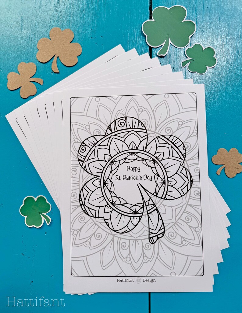 May include: A stack of blank white cards featuring a black and white printable design of a shamrock with the text "Happy St. Patrick's Day" inside the shamrock. The shamrock is surrounded by a mandala design. The cards are on a blue wooden surface with green and brown shamrock cutouts.