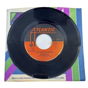 May include: A black vinyl record with the title "I'M COMING HOME" by Spinners, on an Atlantic label. The record is set against a colorful sleeve with stripes of purple, green, and blue. The record is a 45 RPM single.