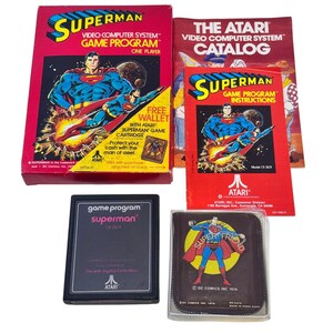 May include: A vintage Atari Superman video game set including a red cardboard box, an instruction manual, a game cartridge, and a brown wallet. The box and manual feature Superman graphics and text. The black cartridge is labeled Superman CX-2631.