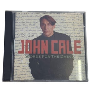 May include: A compact disc case featuring the album Words for the Dying by John Cale. The cover art displays a portrait of John Cale in a black jacket against a background of handwritten cursive text. The artist name is in large red letters.