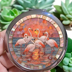 May include: A circular silver compact mirror with a mosaic design. The mosaic depicts pink flamingos in a sunset scene, with orange, red, and pink hues. The flamingos are standing in water, creating a vibrant and detailed image.