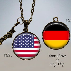 Op de afbeelding: Een ketting met een omkeerbare hanger met aan de ene kant de Amerikaanse vlag en aan de andere kant de Duitse vlag. De hanger is bevestigd aan een bronzen ketting met een klein Vrijheidsbeeld-bedeltje. "Zijde 1" en "Zijde 2" staan op de afbeelding, samen met "Uw keuze van elke vlag".