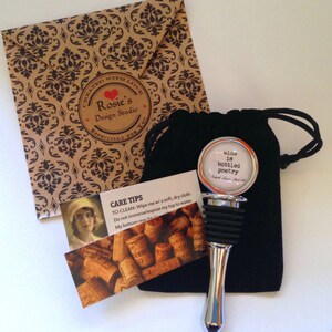 May include: A silver wine bottle stopper with a black rubber seal and a round, silver top with the text "wine is bottled poetry" in black lettering. The stopper is in a black velvet bag with a drawstring.