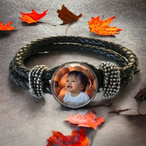May include: A black leather bracelet with a round charm featuring a photo of a young child smiling in a field of pumpkins.