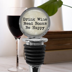 May include: Silver wine bottle stopper with a white and gray patterned top. The top has the text "Drink Wine Read Books Be Happy".