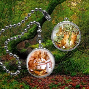 May include: A silver-toned metal pendant necklace featuring a round glass cabochon with a printed illustration of three brown rabbits preparing food, accompanied by a matching circular pendant showing two rabbits picking berries in a garden.