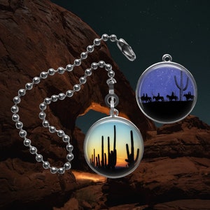 May include: Two round, silver fan pull charms with images of desert landscapes. One charm features a starry night sky with a silhouette of cacti and riders on horseback. The other charm features a sunset with a silhouette of cacti.
