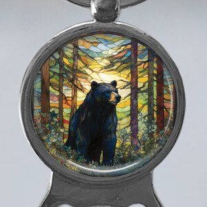 May include: A black bear stands in a woodland setting, surrounded by colourful stained glass trees. The bear is facing right and has a dark brown nose and black eyes. The stained glass background features a variety of colours, including blue, green, yellow, and orange.